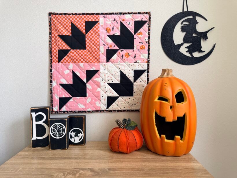 Halloween bat mini quilt displayed on wall above a desk with pumpkins sitting on the desk and a boo sign and witch in a moon picture.